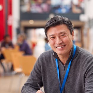Portrait Of Mature Male Teacher Or Student With Digital Tablet Working At Table In College Hall